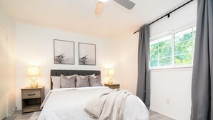 Un dormitorio con una cama, dos mesitas de noche con lámparas, un ventilador de techo, una ventana con cortinas y dos obras de arte colgadas en la pared.