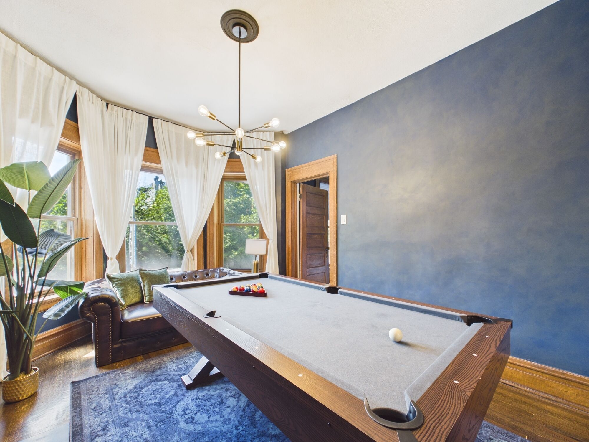 Uma mesa de bilhar em um espaço com uma janela grande, um lustre e um vaso de planta.