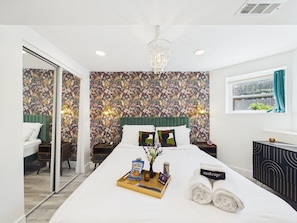 A hotel room with a large bed, a green upholstered headboard, a floral wallpaper, a chandelier, and a view of the outdoors through a window.