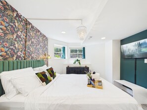 Quarto moderno com uma cama grande, cabeceira verde, parede decorativa, janela com cortinas, cômoda, TV e lustre.