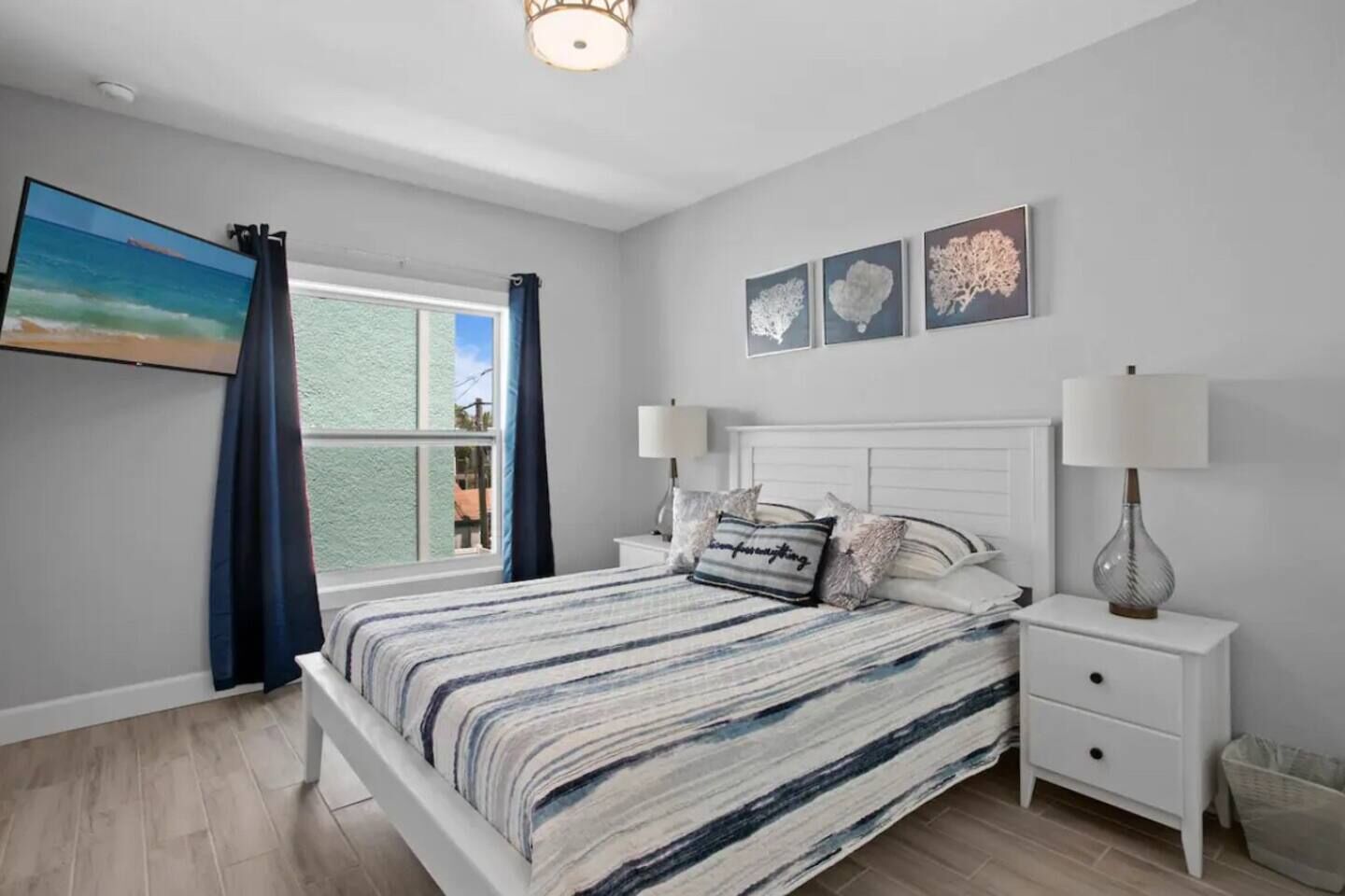 Une chambre à coucher avec un lit, des tables de chevet, une lampe, une fenêtre avec des rideaux et des œuvres d’art accrochées au mur.