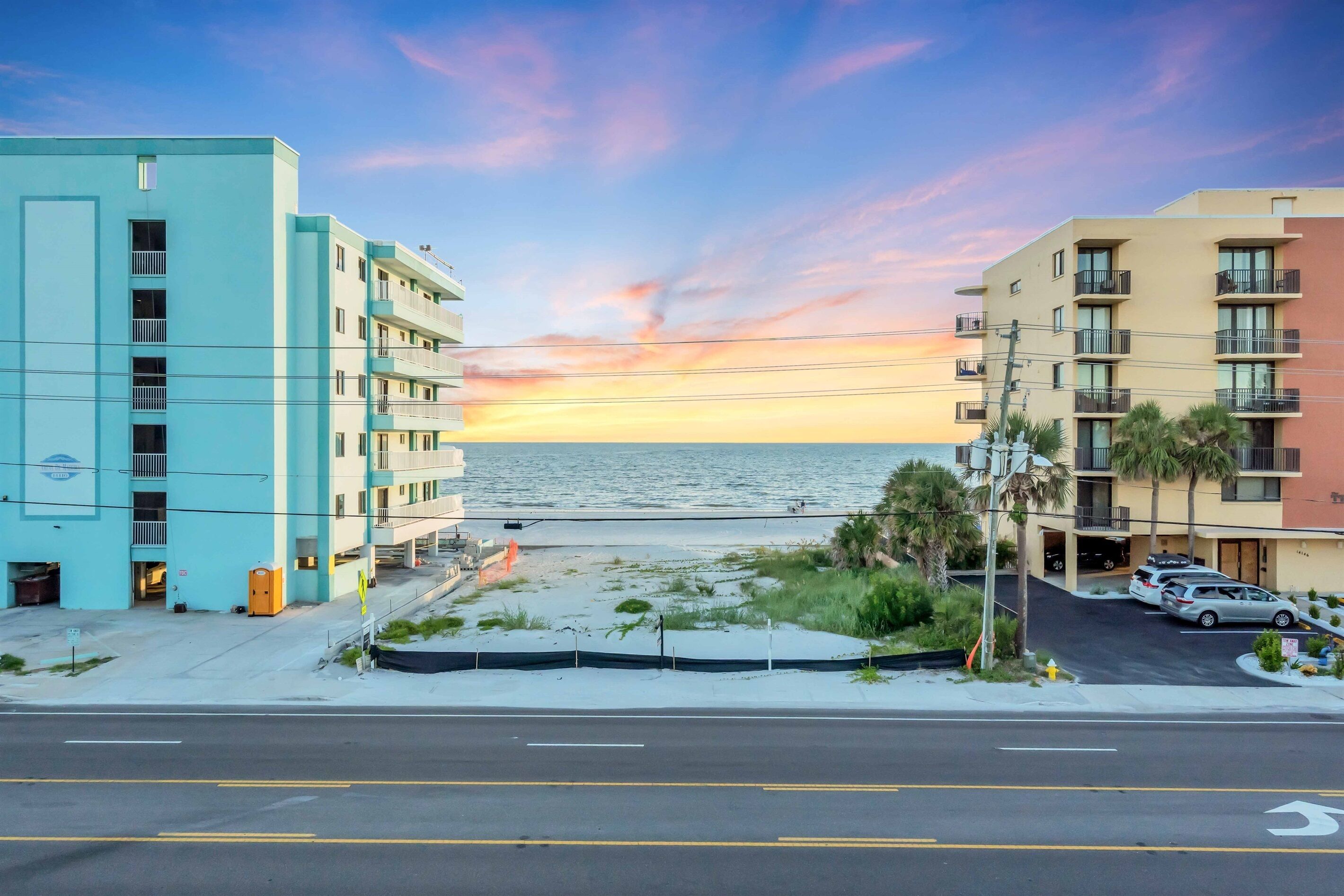 A coastal hotel with a view of the ocean at sunset, featuring multiple balconies and a parking area.