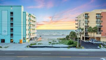 A coastal hotel with a view of the ocean at sunset, featuring multiple balconies and a parking area.