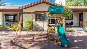 Un parque infantil con tobogán y columpios, una zona de juegos cubierta y una casa con porche al fondo.