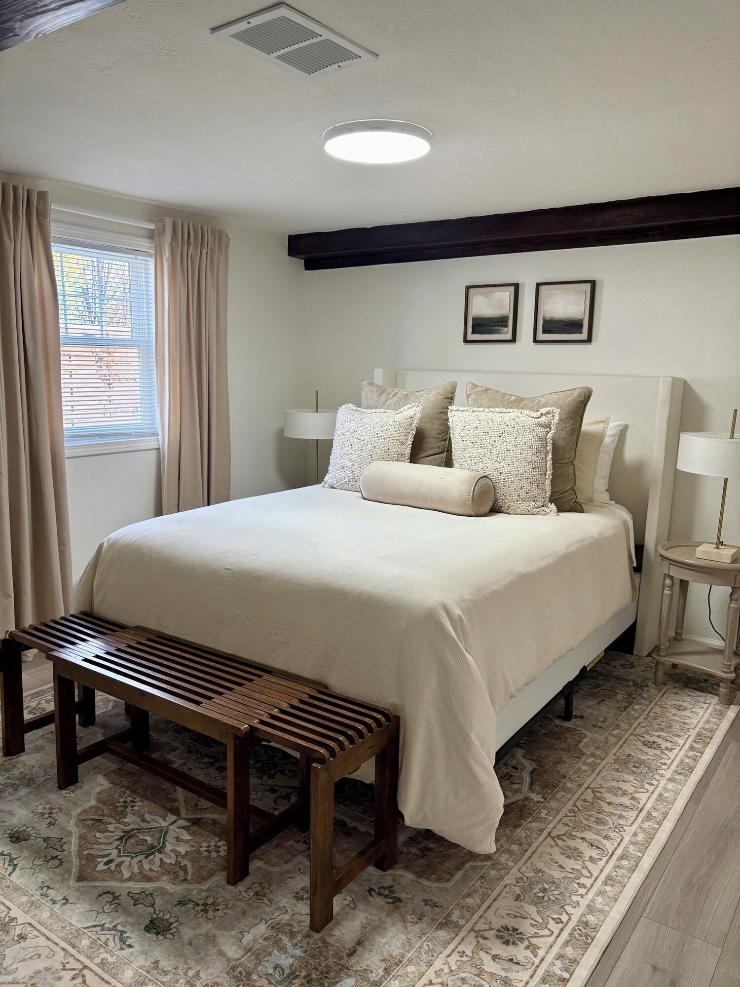 A bedroom with a bed, bedside lamps, a bench, a patterned rug, and a window with curtains.