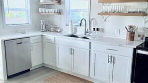 A modern kitchen with white cabinetry, a double sink, and a built-in oven. There are two windows with blinds, a wooden shelf above the sink, and a small rug on the floor.
