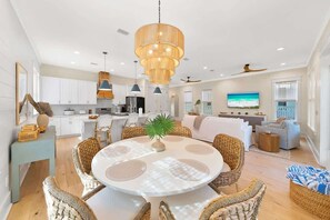 Une cuisine moderne avec des meubles blancs, un coin repas avec des chaises en osier et un grand téléviseur dans le salon.