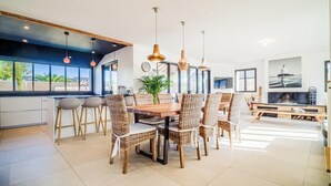 Une cuisine moderne avec un coin repas, comprenant un grand îlot central, un éclairage suspendu et une vue sur l’extérieur.