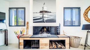 Un salon moderne avec une cheminée, un tableau représentant un phare et une grande fenêtre.