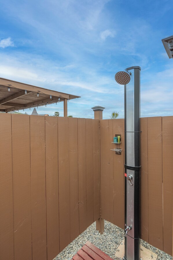 A metal showerhead attached to a vertical pole, with a wooden fence and a small shelf with toiletries in the background. - AIRSTREAM GLAMPING! Jacuzzi, BBQ & Chill! (Valley Center)