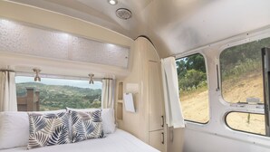 Een slaapkamer met een bed, kussens, een raam en uitzicht op een glooiend landschap.