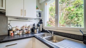 Une cuisine avec des meubles blancs, un plan de travail noir, un évier à deux bacs et une fenêtre donnant sur les arbres.