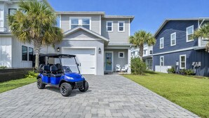 A residential area with a paved driveway, a blue golf cart, and houses with a light gray and dark blue exterior. - Destin/Miramar 4BR/4BA | Golf Cart | Pool | 1st Floor BR | Pets OK | Fenced Yard (Miramar Beach)