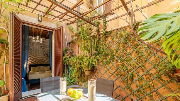 A glass table with a bowl of fruit and two glasses of a white beverage, a bed with blue curtains, and a wooden pergola with plants. - One Bedroom Apartment (ROMA)