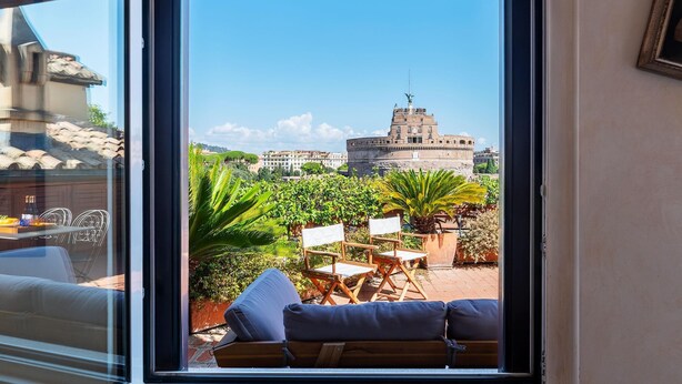 Une vue depuis une fenêtre donnant sur une cour intérieure avec un bâtiment en pierre, du mobilier d’extérieur et de la verdure.