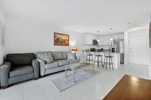 A modern living room with a grey sofa, a coffee table, and a kitchen area with white cabinets and a microwave. - OV 222 Tennis Court View - Welcome To Paradise! (Fort Pierce)