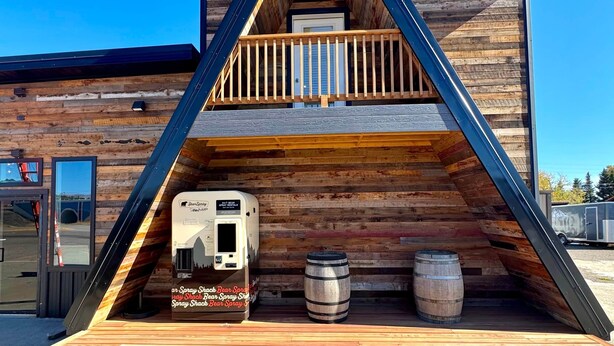 Une cabane avec un toit en pente, un balcon et une terrasse comprenant un camping-car et des barils.