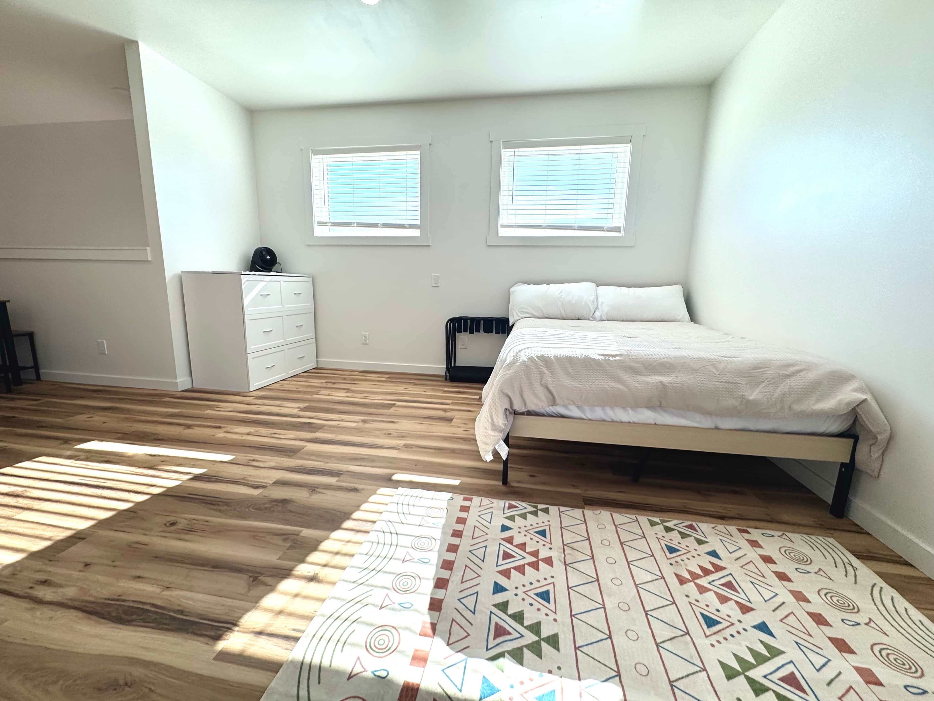 Une chambre à coucher avec un lit, deux fenêtres, une commode et un tapis à motifs.