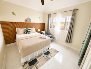 Un dormitorio con cama, mesita de noche, lámpara, silla y vistas a través de una ventana.