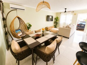 A dining area with a round table, wicker chairs, a hanging chair, and a TV on the wall.