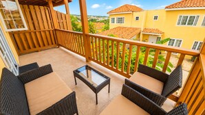A balcony with wicker furniture, a glass table, and a view of yellow buildings and greenery. - NEW! Hilltop Escape with Ocean Views (Montego Bay)