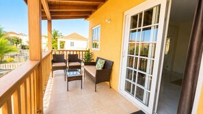 A covered balcony with wicker furniture, a glass table, and a sliding glass door leading to an interior space.