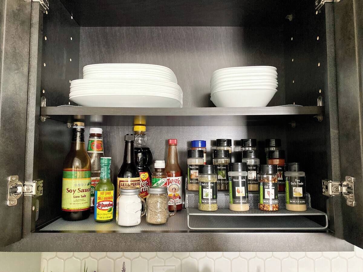A kitchen cabinet with shelves holding white plates, bottles of soy sauce, hot sauce, and various spices.