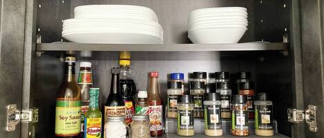 A kitchen cabinet with shelves holding white plates, bottles of soy sauce, hot sauce, and various spices.