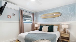 Una camera da letto ordinata con un letto, comodini, una tavola da surf appesa al muro e una finestra con tende.