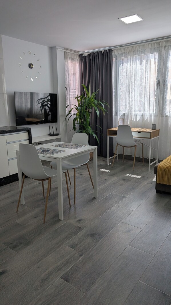 A modern living room with a dining table, chairs, a TV, a plant, and a window with curtains.