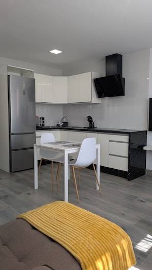 Cozinha moderna com armários brancos, geladeira de aço inoxidável, coifa preta, mesa de jantar branca com duas cadeiras e um cobertor amarelo no chão.