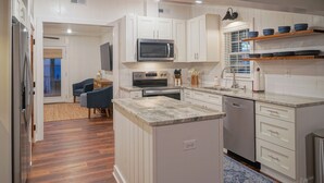 Una cocina moderna con armarios blancos, microondas integrado, horno y lavavajillas. Hay un comedor con una silla azul y una mesita.