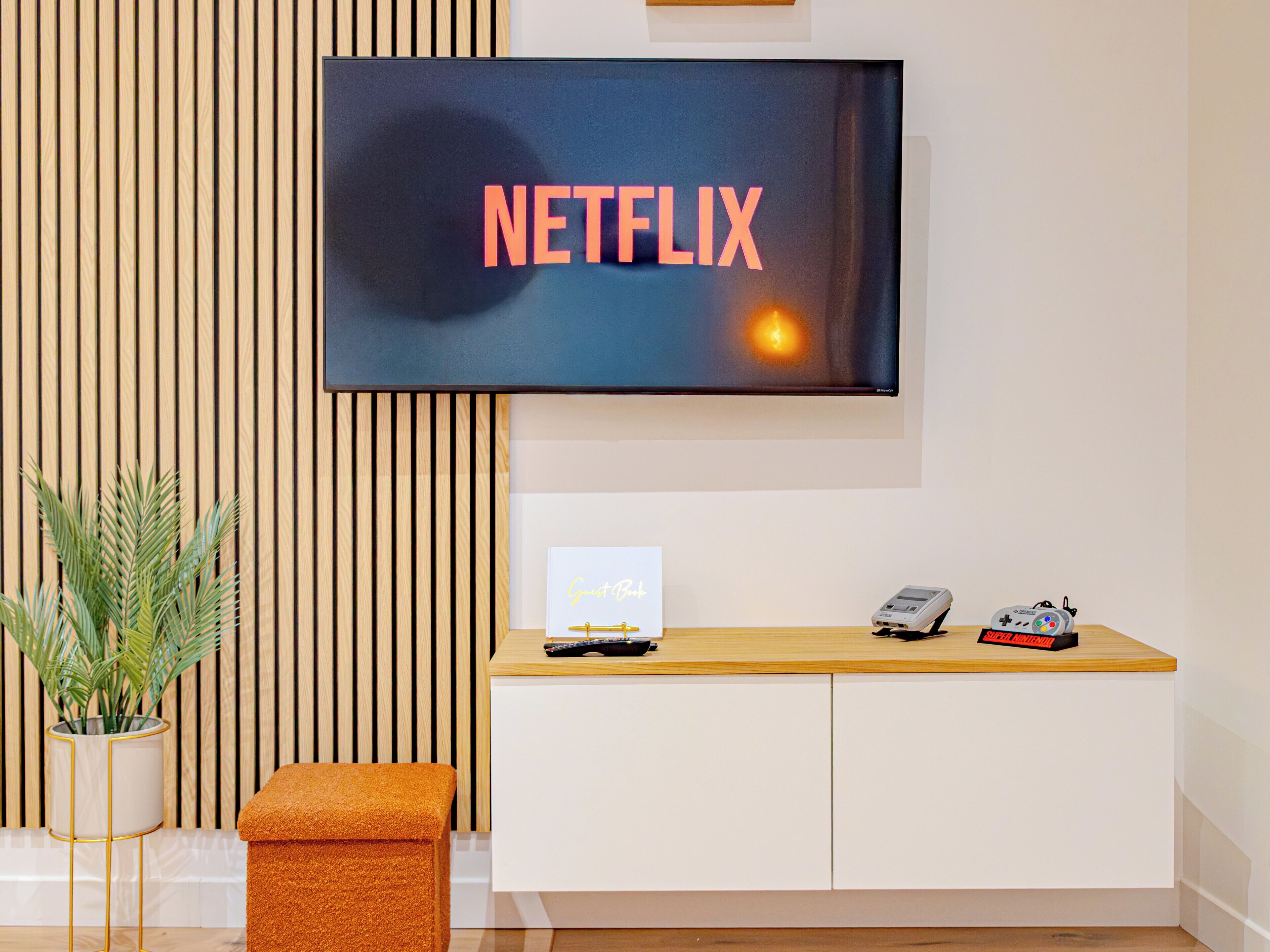 A modern living room with a flat-screen TV displaying 'NETFLIX' on the screen, a small table with a remote control, a plant, and a bench.