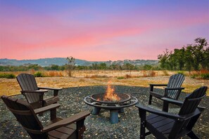 A fire pit with four Adirondack chairs arranged around it, set on a gravel surface with a view of a hilly landscape in the background.