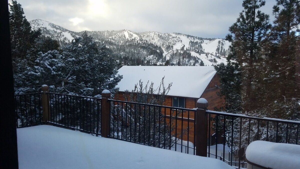 A wooden cabin with a snow-covered roof, surrounded by snow-laden trees and mountains in the background.