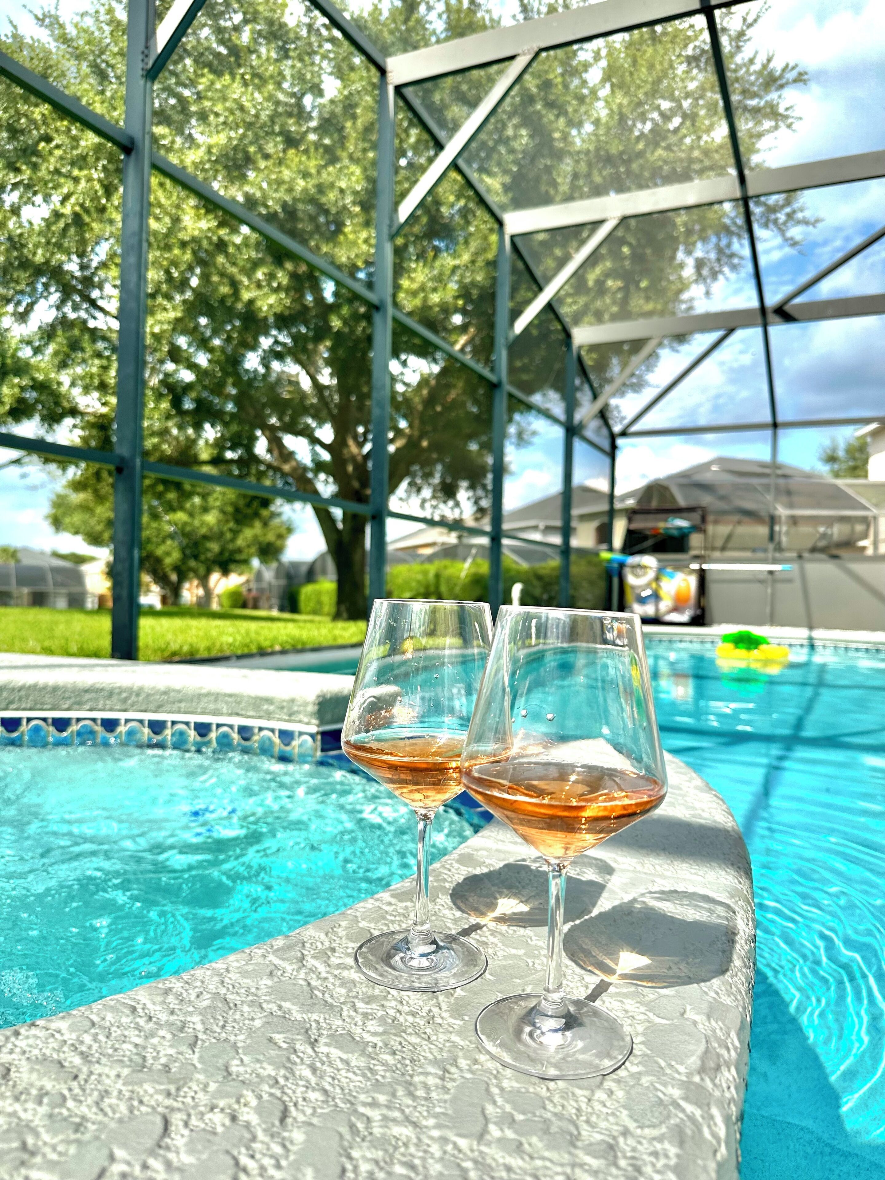Two wine glasses on a poolside ledge with a clear blue pool and a covered area in the background.