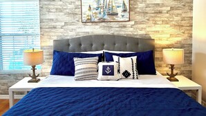 A bedroom with a grey upholstered headboard, a blue quilted bedspread, and matching pillows. A nautical-themed painting hangs above the bed, and there are two bedside lamps on white nightstands.