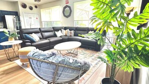 A modern living room with a sectional sofa, a round coffee table, and a wicker chair. There is a large potted plant and a small table with a lamp.