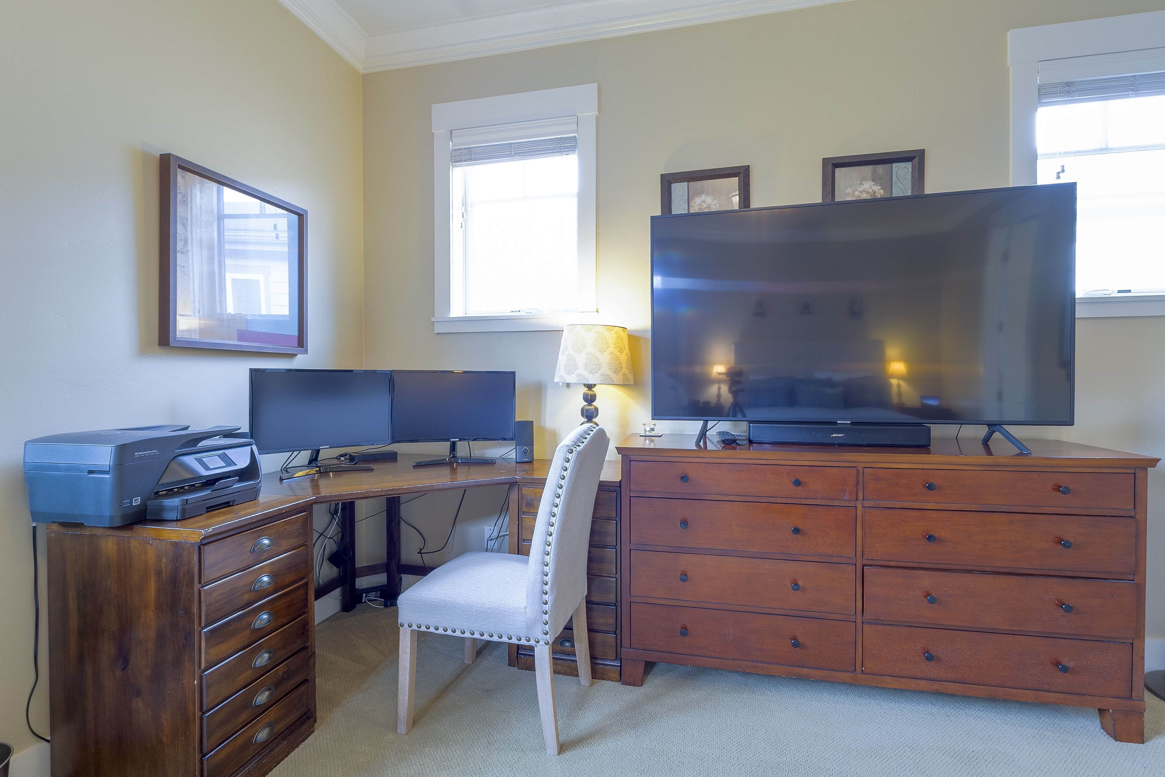 Een werkplek aan huis met een bureau, twee monitoren, een printer, een televisie, een stoel en een houten dressoir.