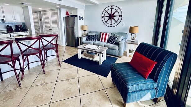 A modern living room with a sofa, coffee table, and chairs. There is a kitchen area with cabinets and a refrigerator. A large window provides natural light. - Islands Winds East - Unit 506 (Gulf Shores)
