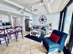 A modern living room with a sofa, coffee table, and chairs. There is a kitchen area with cabinets and a refrigerator. A large window provides natural light.