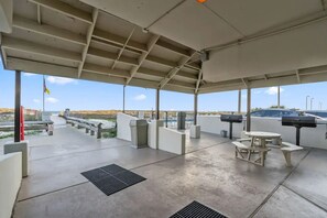 A covered outdoor area with picnic tables and a barbecue grill, overlooking a beach and a road.