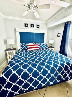 A bedroom with a blue patterned bedspread, a white ceiling fan, and a nightstand with a lamp.