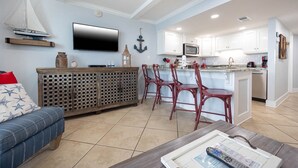 A modern living room with a sofa, a coffee table, and a TV. There is a kitchen area with white cabinets and a microwave. - Islands Winds East - Unit 506 (Gulf Shores)