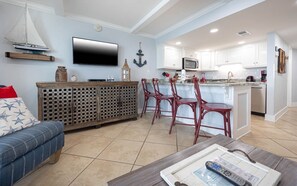 Ein modernes Wohnzimmer mit Sofa, Couchtisch und Fernseher. Es gibt eine Küchenzeile mit weißen Schränken und einer Mikrowelle.