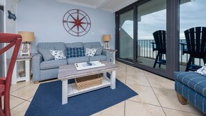 A cozy living area with a grey sofa, two side tables with lamps, a wooden coffee table, and a large window with a view of the ocean. - Islands Winds East - Unit 506 (Gulf Shores)