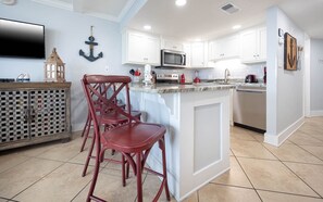 A modern kitchen with white cabinetry, a central island with bar stools, a flat-screen TV, and nautical-themed decor.