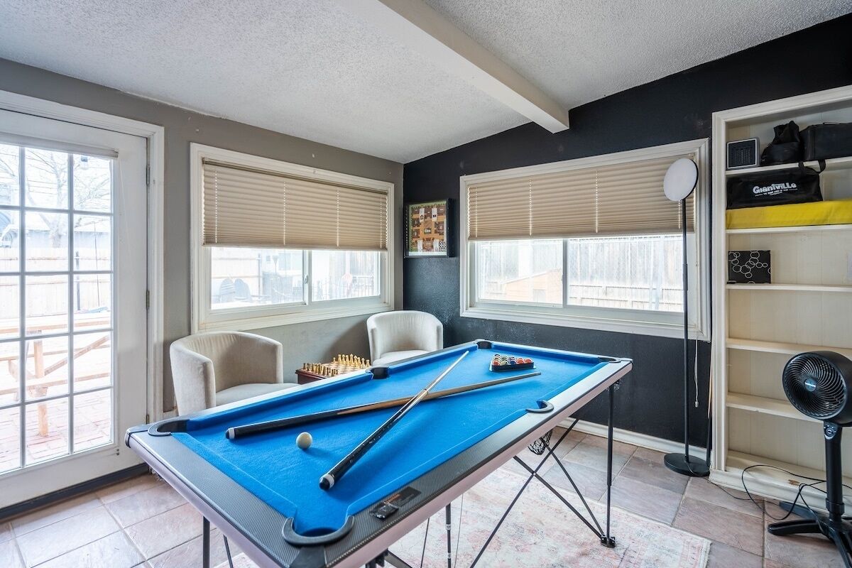 Une table de billard dans une pièce dotée de grandes fenêtres, d’une chaise et d’un ventilateur.