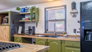 Eine Küche mit Gasherd, Spüle und Kühlschrank. Es gibt grüne Schränke, ein Fenster mit Jalousien sowie ein Regal mit Pflanzen und Küchenutensilien.