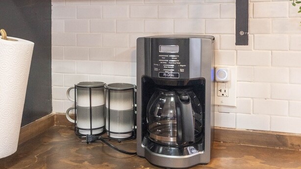 Eine Küche mit Kaffeemaschine, Regalen mit Geschirr und einer hängenden Pflanze.